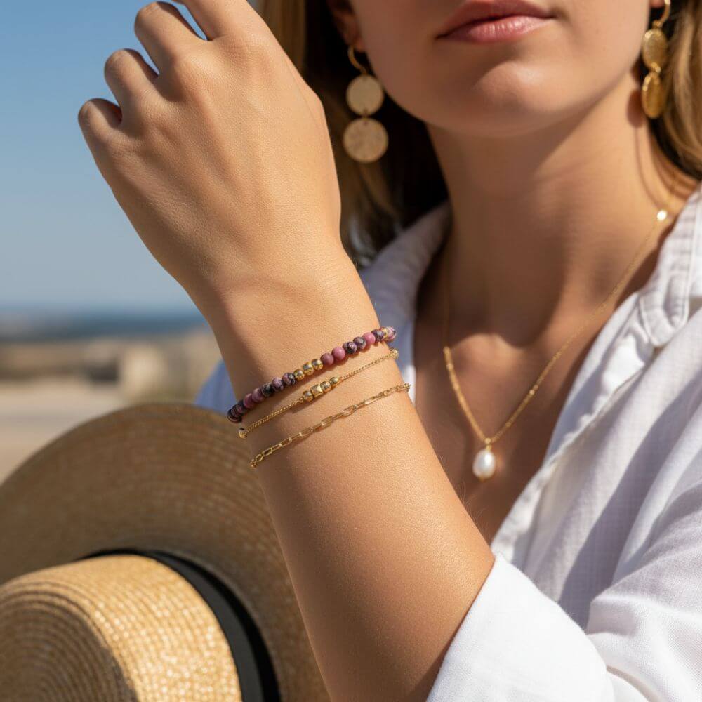 Femme portant une accumulation de bracelets dorés et en perles roses, un collier avec pendentif perle blanche et des boucles d'oreilles pendantes dorées.