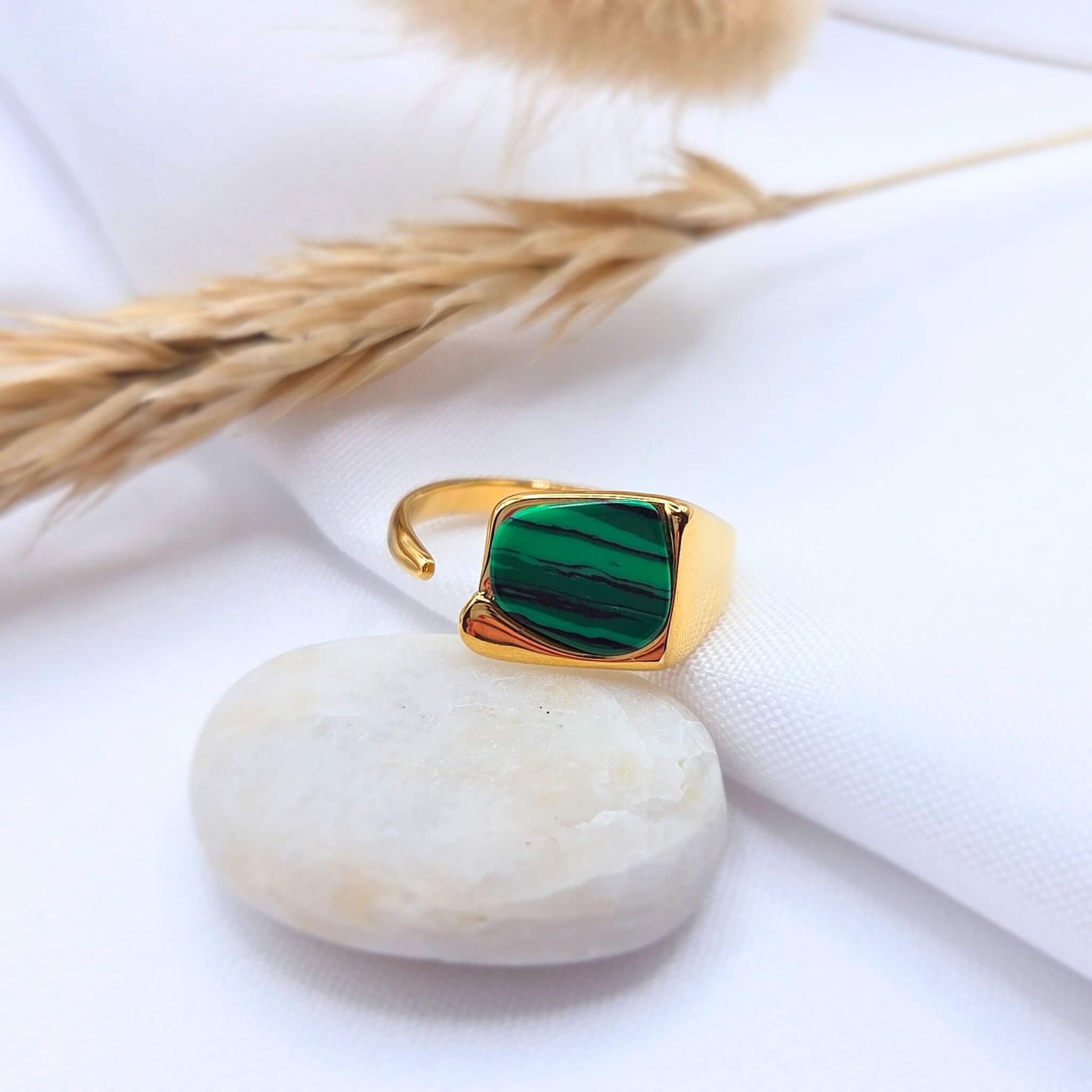 Bague chevalière moderne en acier inoxydable doré avec un plateau rectangulaire aux bords arrondis. Le plateau est serti d'une pierre naturelle de Malachite d'un vert profond présentant des strates horizontales noires. Le bijou, doté d'un anneau ouvert et ajustable, est posé sur un galet blanc devant un arrière-plan composé d'un tissu blanc et de graminées séchées.