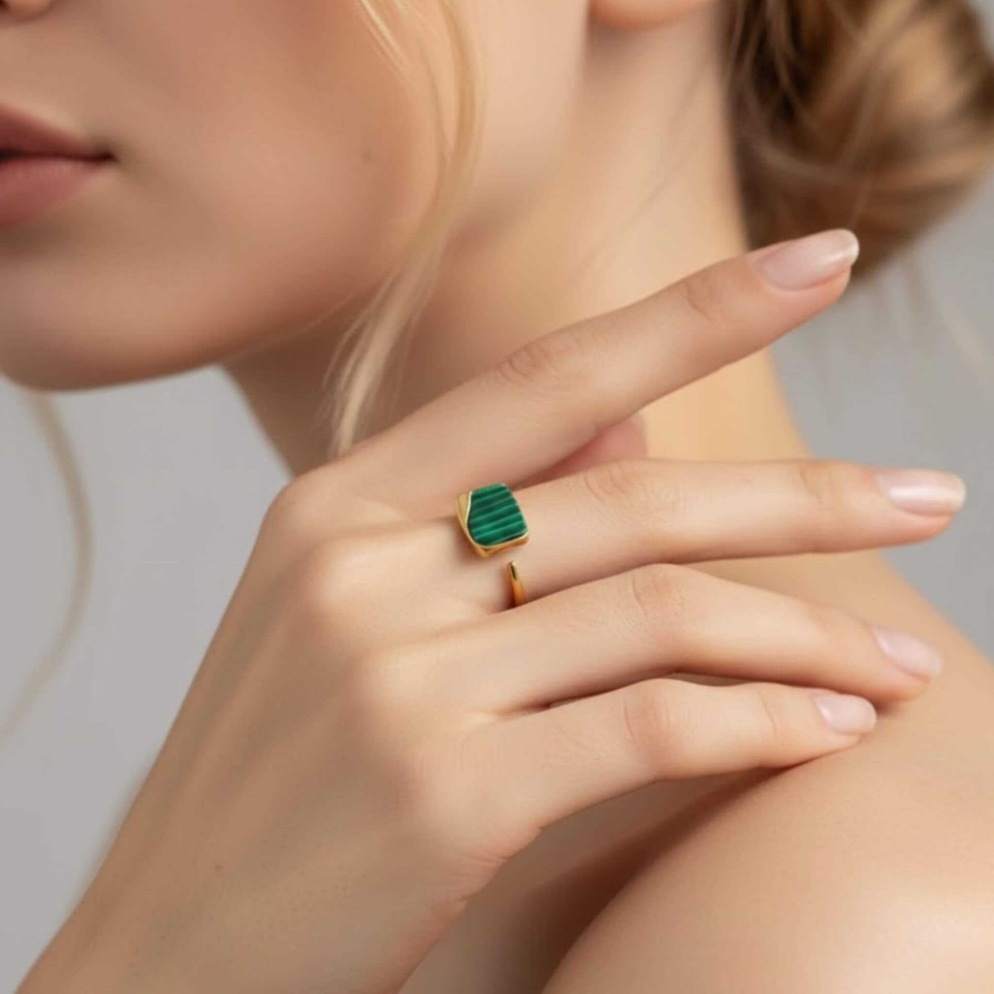 Gros plan sur la main d'une femme posée sur son épaule, mettant en valeur une bague moderne en plaqué or ornée d'une pierre rectangulaire en malachite vert vibrant aux rainures caractéristiques. La bague présente un anneau ouvert au design minimaliste.