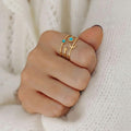 Gros plan d'une main portant la bague dorée aux motifs célestes sur l'index. Les détails du bijou, notamment les deux pierres turquoises et les formes de lune et d'étoile, se détachent sur le fond doux d'un pull en laine blanche à mailles aérées. La lumière douce met en valeur le fini mat de l'or et l'éclat bleu des pierres.