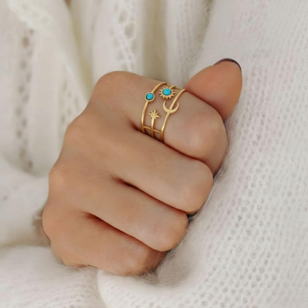 Gros plan d'une main portant la bague dorée aux motifs célestes sur l'index. Les détails du bijou, notamment les deux pierres turquoises et les formes de lune et d'étoile, se détachent sur le fond doux d'un pull en laine blanche à mailles aérées. La lumière douce met en valeur le fini mat de l'or et l'éclat bleu des pierres.