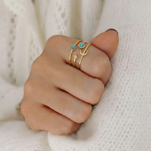 Gros plan d'une main portant la bague dorée aux motifs célestes sur l'index. Les détails du bijou, notamment les deux pierres turquoises et les formes de lune et d'étoile, se détachent sur le fond doux d'un pull en laine blanche à mailles aérées. La lumière douce met en valeur le fini mat de l'or et l'éclat bleu des pierres.