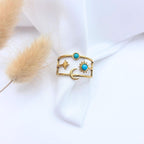 Photographie en gros plan d'une bague dorée au style bohème, présentée sur un tissu blanc texturé. La bague est large et composée de trois anneaux horizontaux reliés entre eux. Elle est ornée de motifs célestes : une étoile polaire, un croissant de lune et un soleil serti d'une pierre turquoise ronde. Une deuxième pierre turquoise est fixée sur l'anneau supérieur. À gauche, on aperçoit le flou délicat d'une plante séchée de type sétaire.