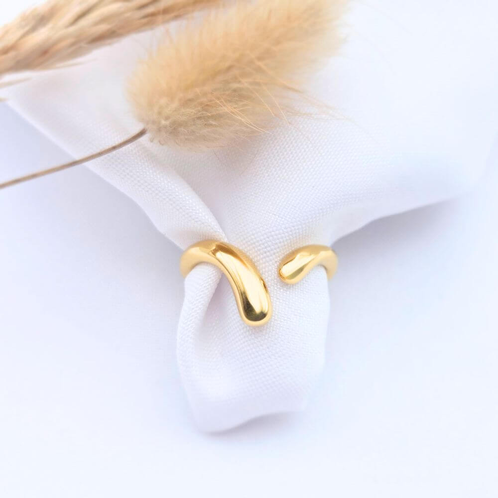 Bague NAMI en acier inoxydable doré au design de vague organique et minimaliste, posée sur un tissu blanc avec un épi de blé séché.