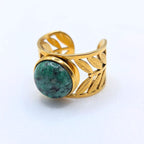 Gros plan d'une large bague ajustable en acier inoxydable doré, de type bandeau, sur un fond blanc épuré. L'anneau présente un travail de ciselure délicat avec des motifs de feuilles ajourées. Au centre, le bijou est serti d'une pierre naturelle de Turquoise africaine ronde bombée (cabochon) d'un vert forêt moucheté de noir.