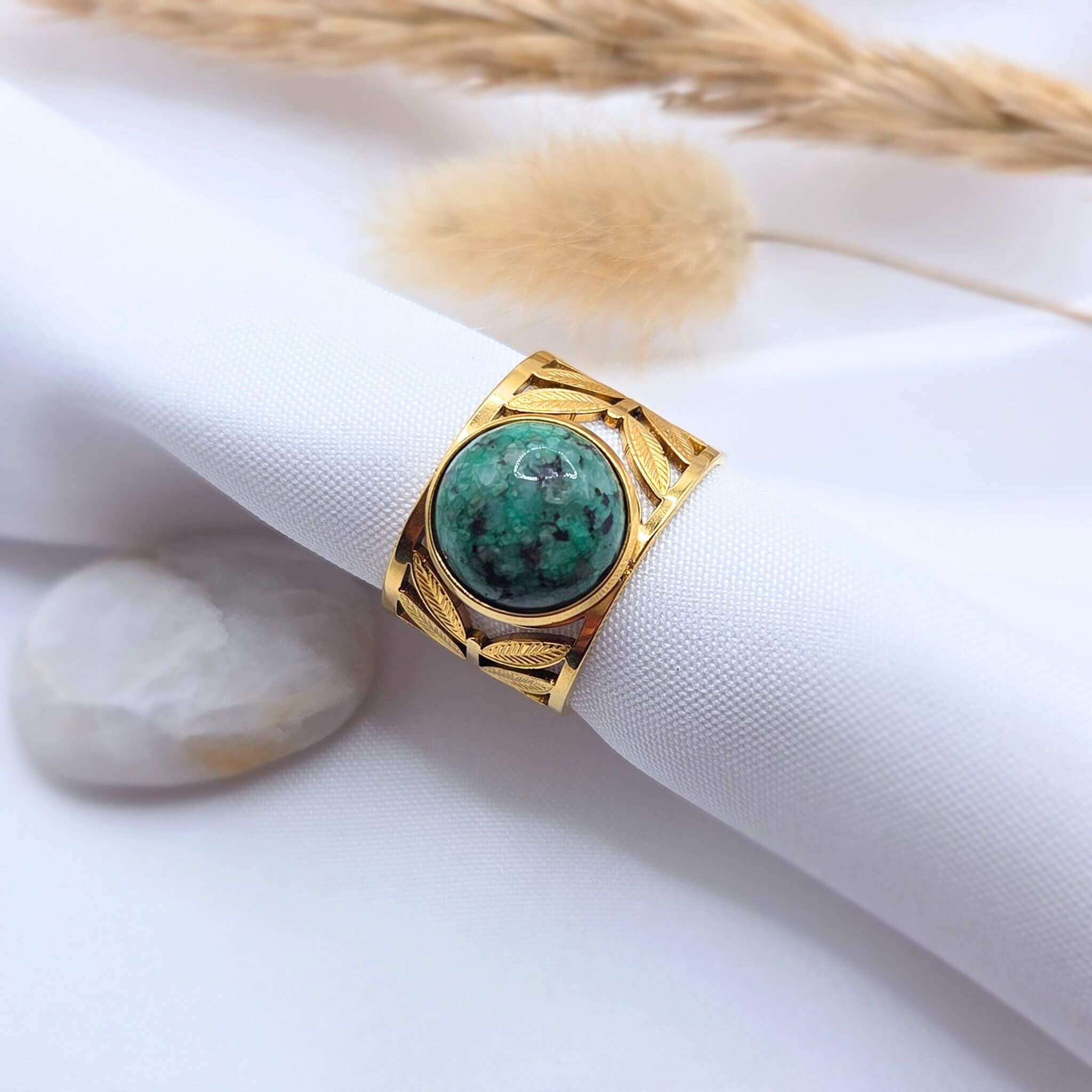 Bague bandeau en métal doré avec des motifs de feuilles ciselées, enroulée autour d'un support en tissu blanc cylindrique. Le bijou est orné d'une pierre de Turquoise Africaine ronde bombée d'un vert forêt intense avec des inclusions foncées. En arrière-plan, un décor doux composé d'un galet blanc et de graminées séchées.