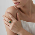 Gros plan d'une femme portant une large bague bandeau en acier inoxydable doré sur son index. L'anneau est décoré de motifs de feuilles ciselées et serti d'une pierre ronde de turquoise africaine d'un vert forêt profond. La main est posée délicatement sur son épaule, devant un vêtement blanc plissé.