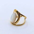 Vue latérale d'une bague large en acier inoxydable plaqué or 18 carat brillant, présentant un design épuré avec des anneaux incurvés. Au centre, une pierre naturelle ovale de jade blanche lisse est sertie dans une monture ouverte. L'ensemble est photographié sur un fond blanc uni, mettant en relief les courbes du bijou.