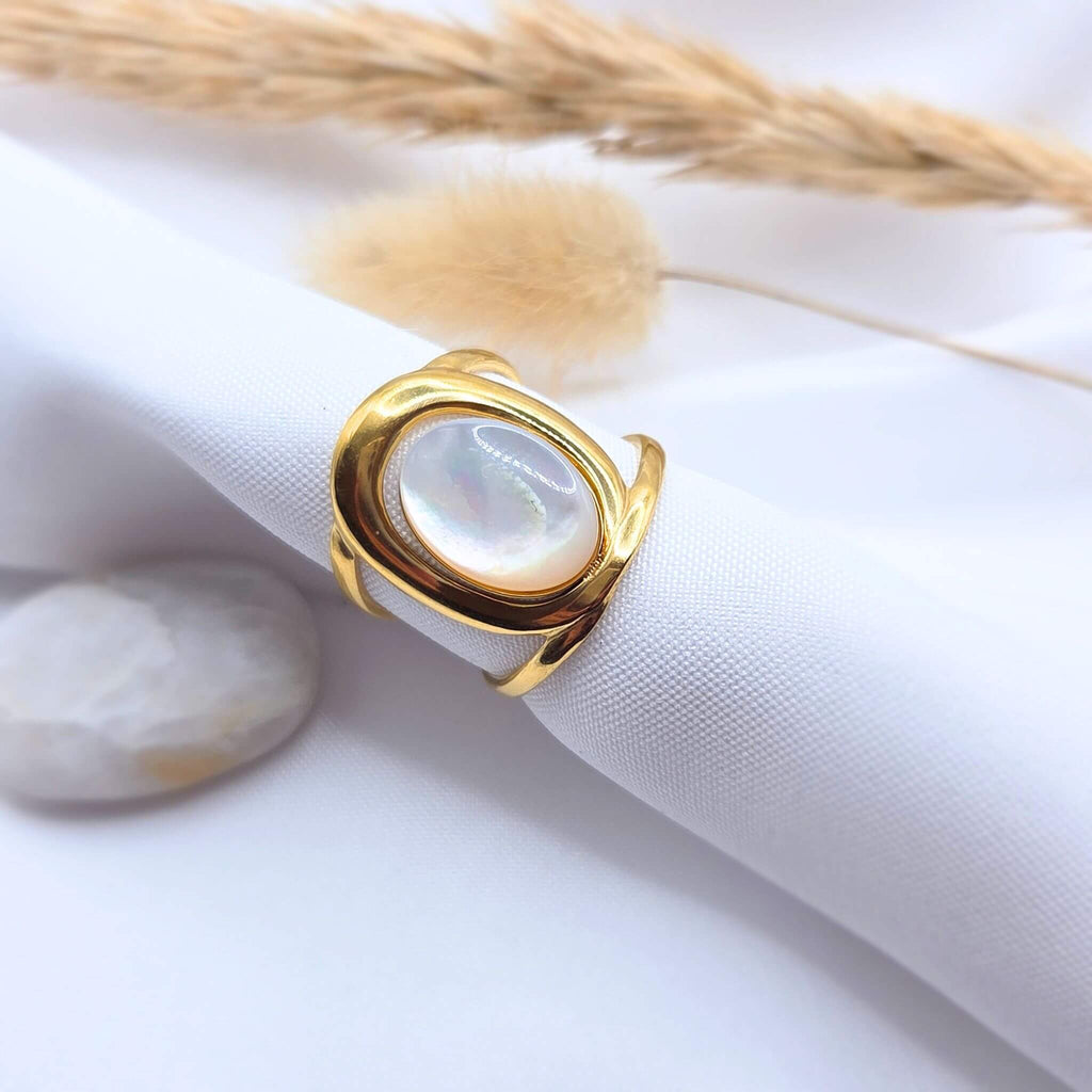 Bague plaqué or 18 carat moderne avec une pierre ovale en jade blanche irisée, fixée sur un support cylindrique en tissu blanc. La mise en scène inclut un galet poli et un plumeau de pampa séchée en arrière-plan, créant une ambiance douce et lumineuse.