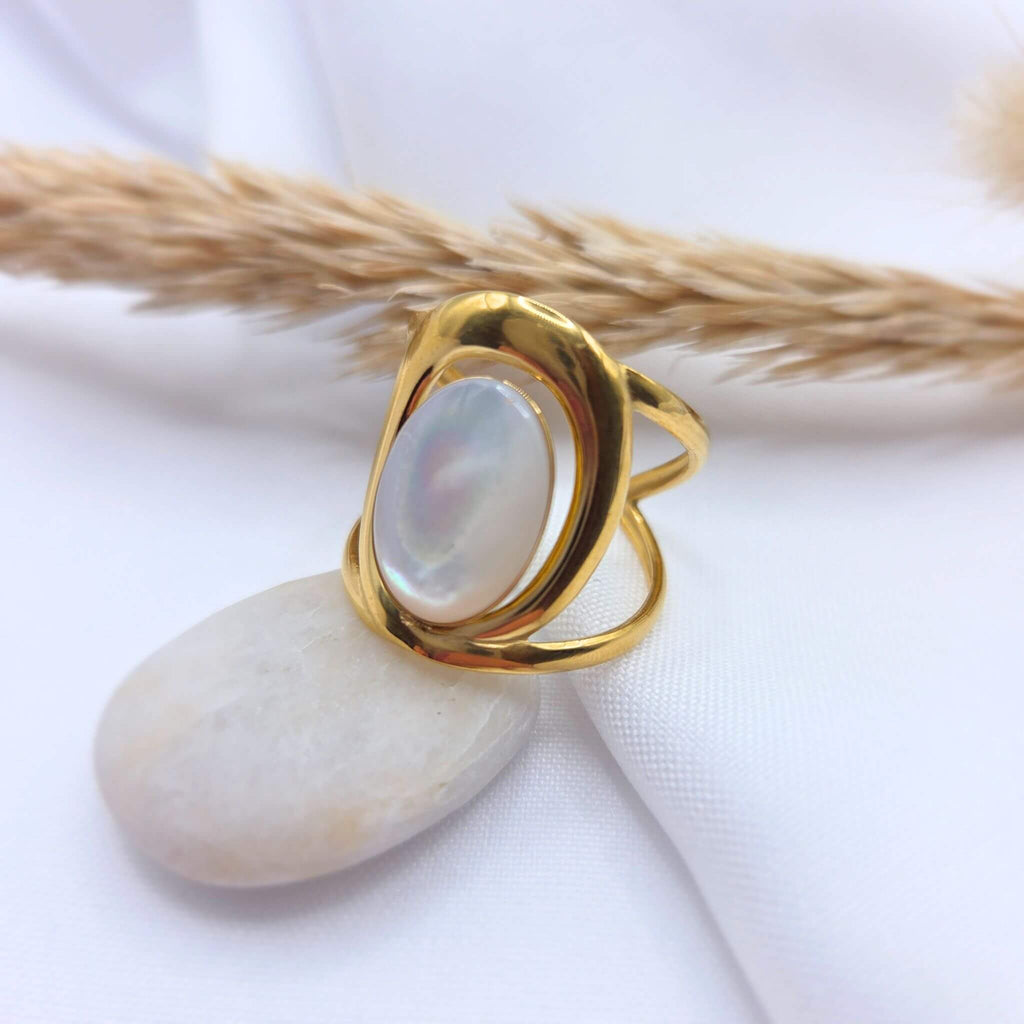 Bague en acier inoxydable doré au design moderne et incurvé, sertie d'une pierre  ovale en jade blanche aux reflets irisés. Le bijou est posé en équilibre sur un galet blanc lisse, avec un arrière-plan composé d'un tissu blanc drapé et d'un épi de graminée séchée.