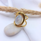 Bague en acier inoxydable doré au design moderne et incurvé, sertie d'une pierre  ovale en jade blanche aux reflets irisés. Le bijou est posé en équilibre sur un galet blanc lisse, avec un arrière-plan composé d'un tissu blanc drapé et d'un épi de graminée séchée.