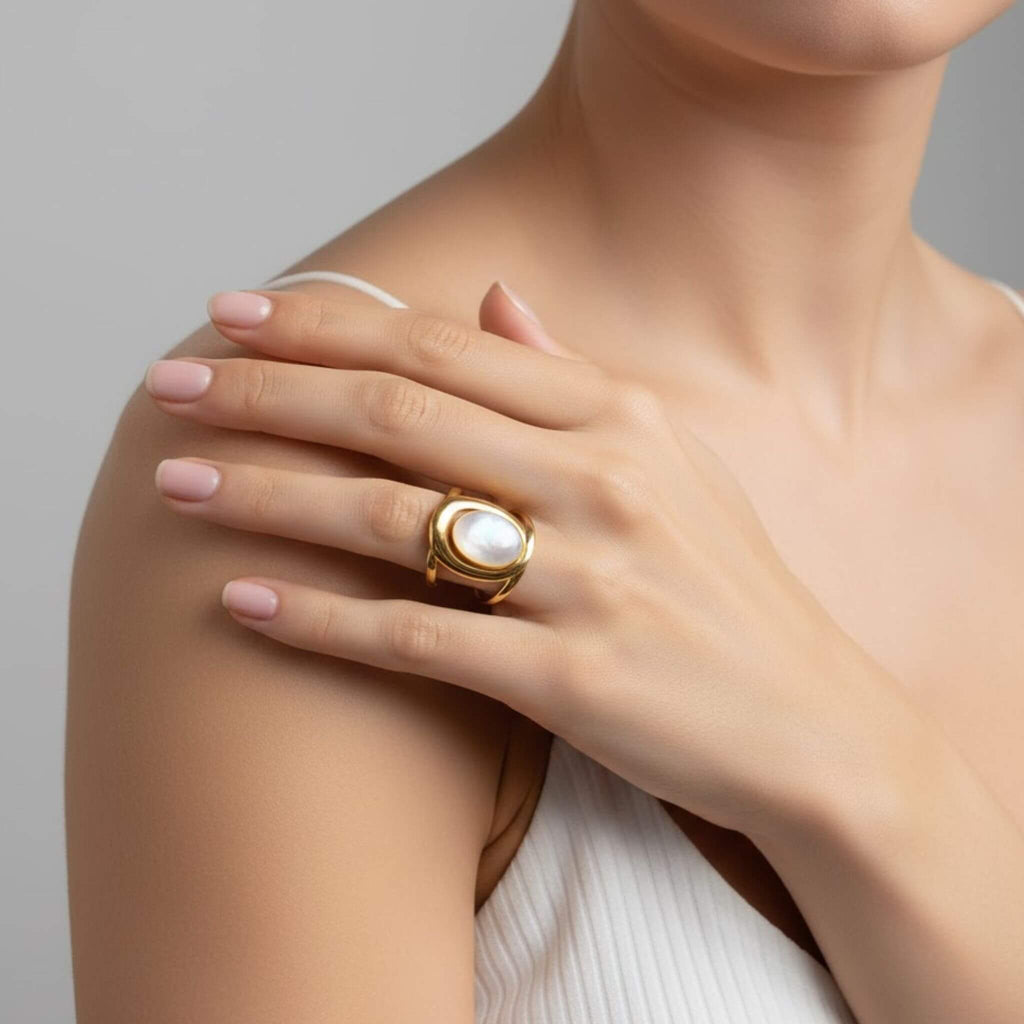Gros plan sur une main de femme posée sur son épaule, arborant une bague dorée imposante sertie d'une pierre de jade blanche ovale. Elle porte un haut blanc à fines bretelles. La lumière douce met en valeur les reflets irisés de la pierre et la finition brillante du métal.