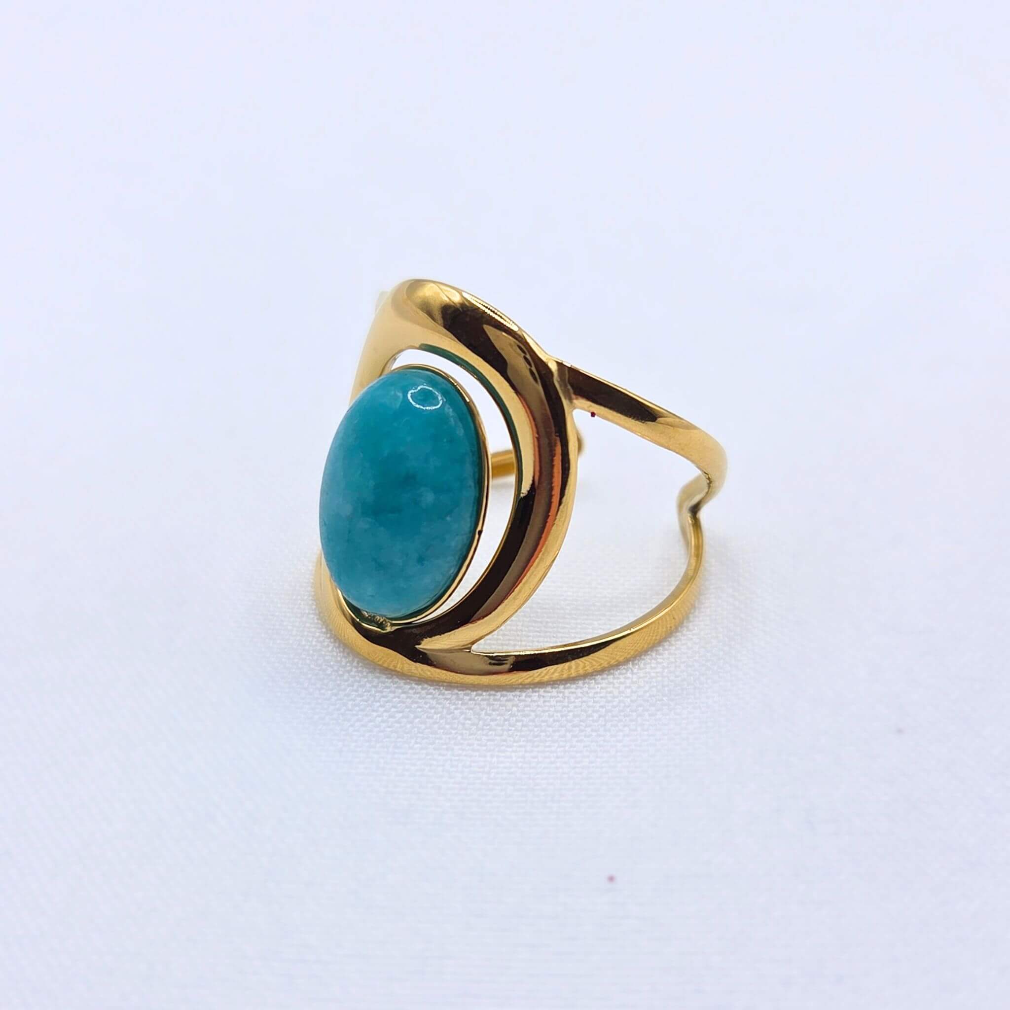 Vue de profil d'une bague imposante en acier inoxydable doré poli, mettant en évidence son design architectural et évidé. Une pierre de jade ovale turquoise est solidement fixée au centre. Le bijou est présenté seul sur un fond blanc texturé épuré.