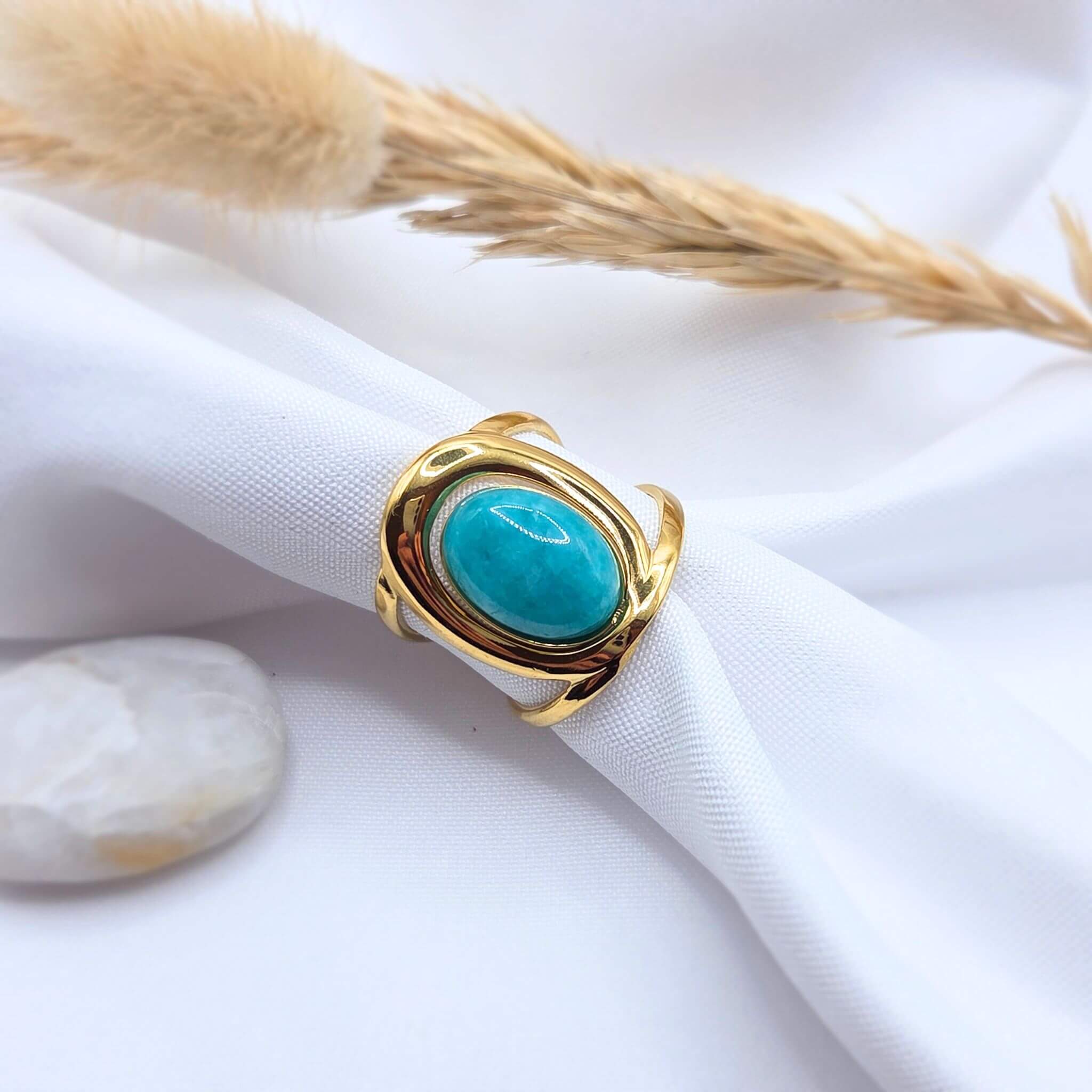 Bague moderne dorée avec une pierre centrale de jade turquoise, présentée sur un tissu blanc drapé. Le design de l'anneau est incurvé et ajouré. Un petit galet blanc et un brin d'herbe séchée décorent l'arrière-plan.