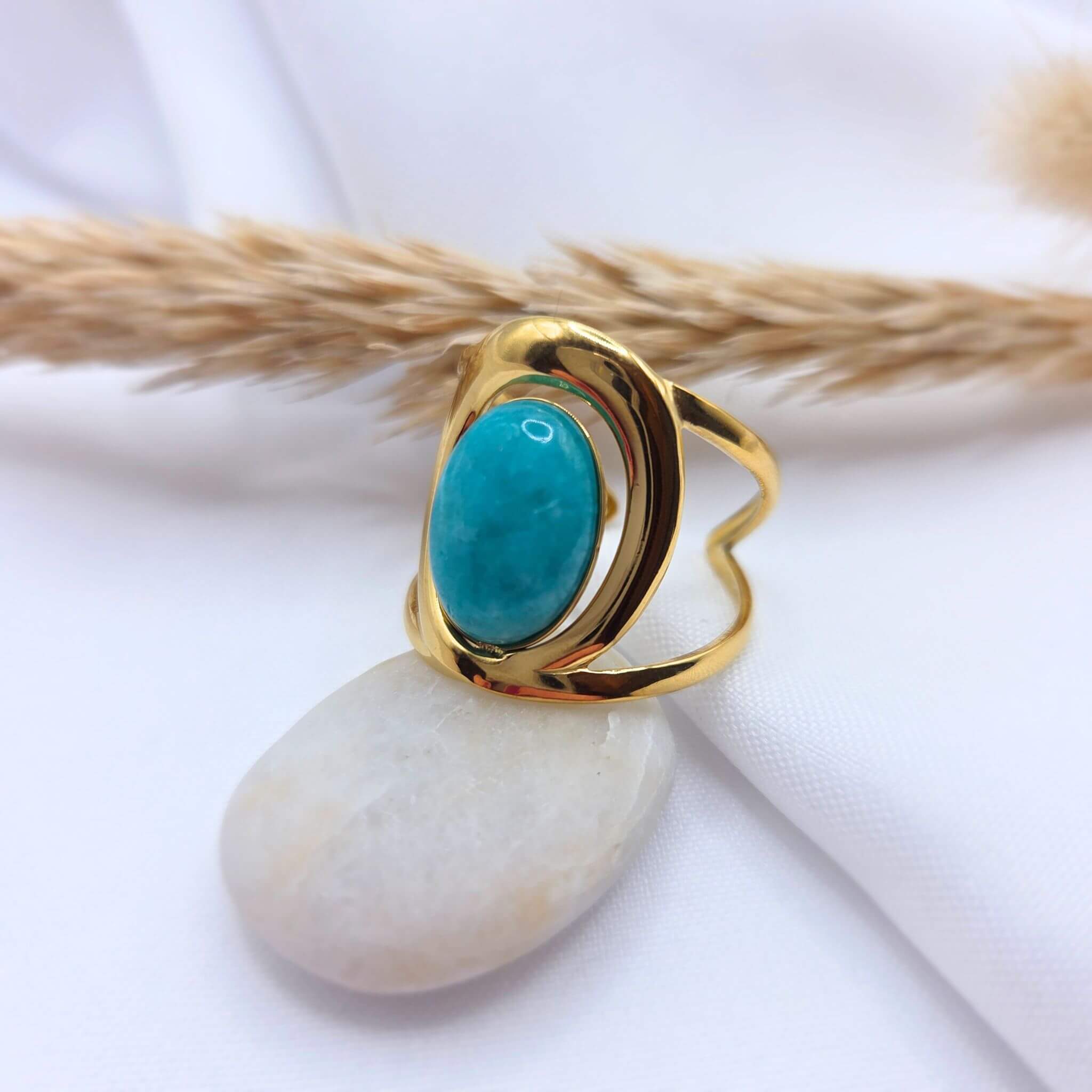 Bague moderne en métal doré ornée d'une pierre ovale de jade de couleur turquoise. L'anneau présente un design incurvé et évidé qui encadre la pierre. Le bijou est posé sur un galet blanc devant un arrière-plan en tissu clair avec un brin d'herbe séchée.