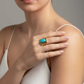 Gros plan sur la main d'une femme posée sur son épaule, portant une bague imposante en or avec une pierre de jade turquoise ovale. La femme porte un haut blanc à fines bretelles. Le style est élégant et lumineux, mettant en valeur le contraste entre le métal doré et la couleur vive de la pierre.