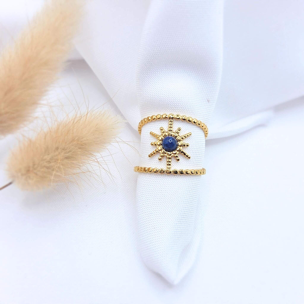 Bague large en acier doré composée de deux anneaux perlés reliés par un motif central en forme de soleil. Le centre du soleil est serti d'une pierre naturelle ronde d'un bleu profond (lapis-lazuli). Le bijou est présenté sur un tissu blanc avec des plumeaux de pampa séchés.