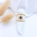 Bague large en acier doré composée de deux anneaux perlés reliés par un motif central en forme de soleil. Le centre du soleil est serti d'une pierre naturelle ronde d'un bleu profond (lapis-lazuli). Le bijou est présenté sur un tissu blanc avec des plumeaux de pampa séchés.