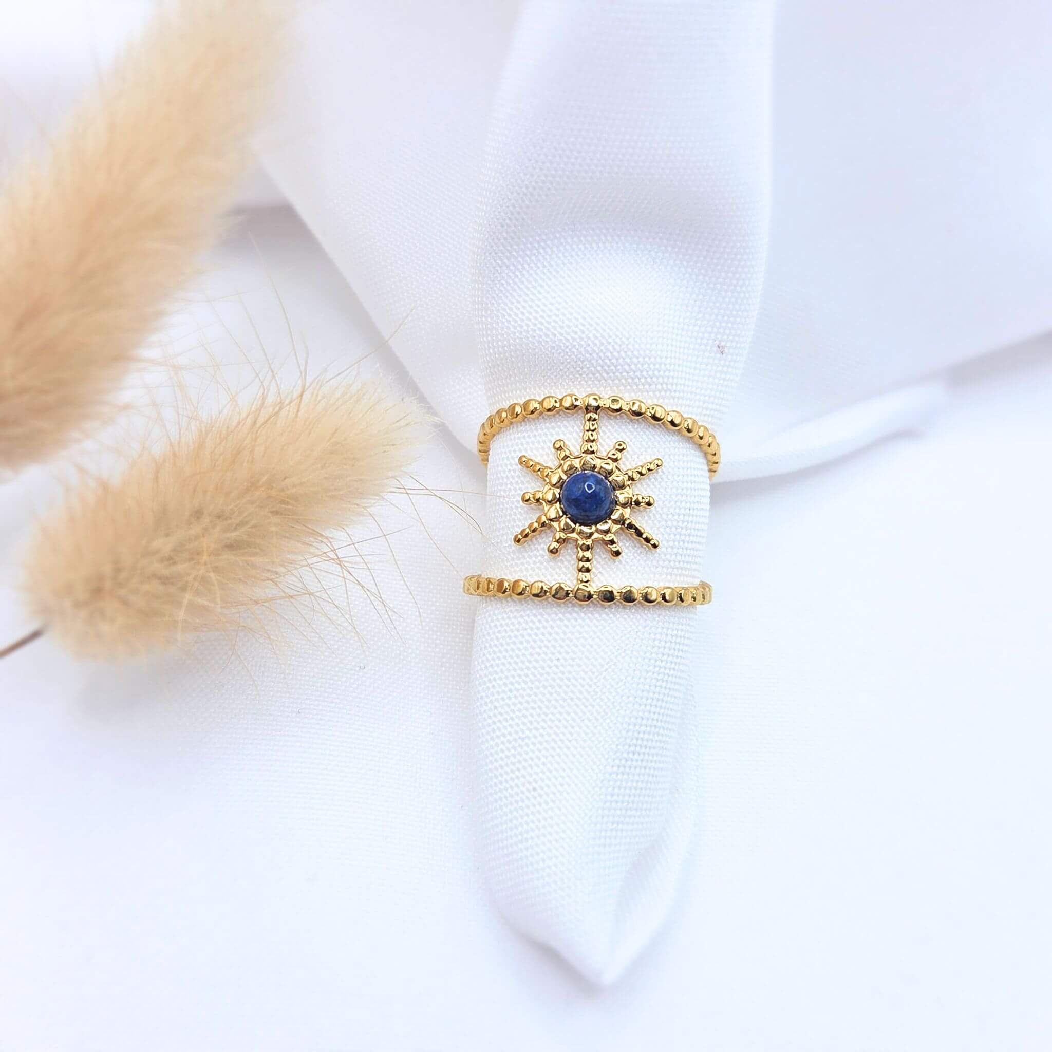 Bague large en acier doré composée de deux anneaux perlés reliés par un motif central en forme de soleil. Le centre du soleil est serti d'une pierre naturelle ronde d'un bleu profond (lapis-lazuli). Le bijou est présenté sur un tissu blanc avec des plumeaux de pampa séchés.