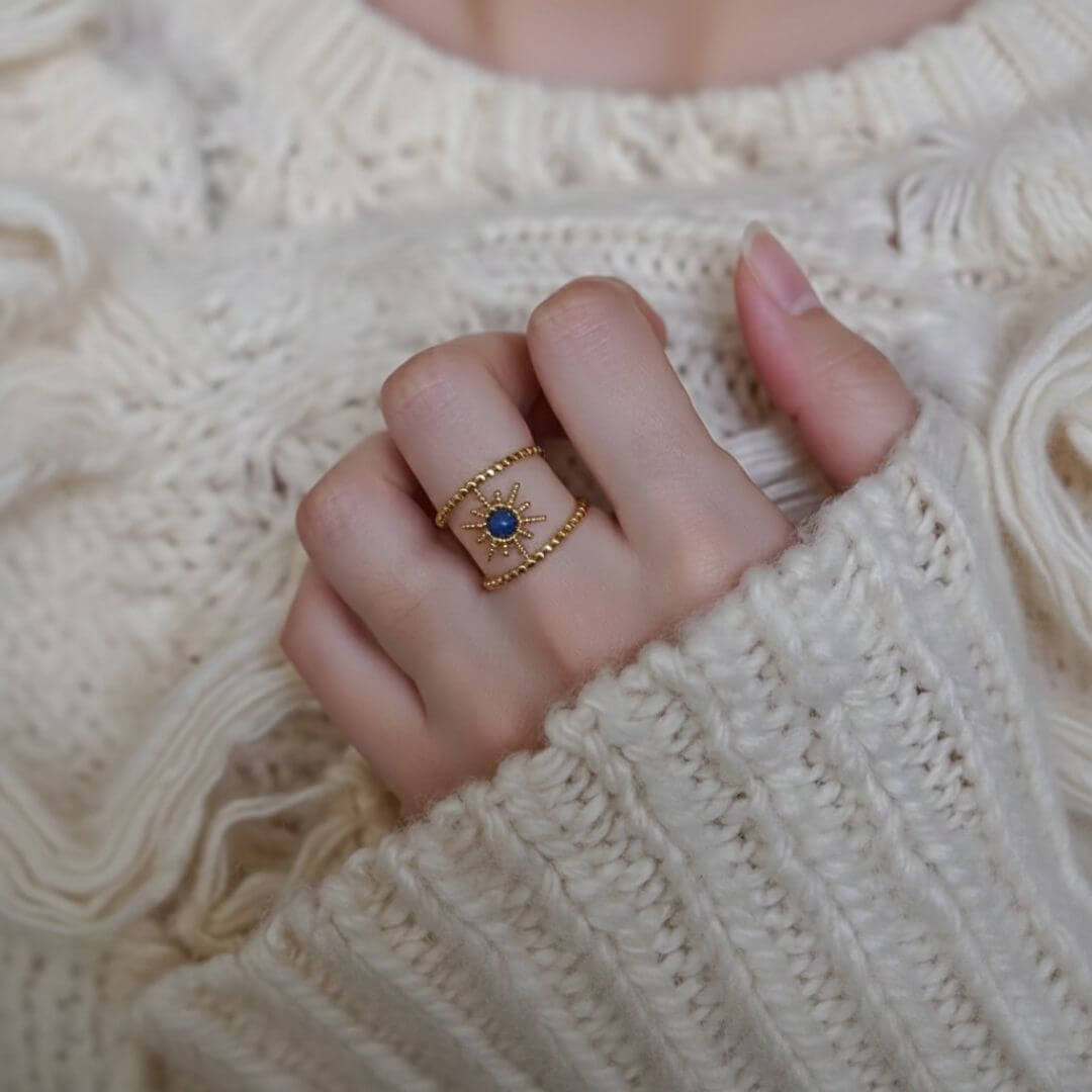 Vue rapprochée de la main d'une personne portant une bague dorée au design céleste sur l'annulaire. La bague se compose de deux anneaux fins à texture perlée, reliés par un motif central en forme de soleil ou d'étoile orné d'une petite pierre bleue au milieu. La personne porte un pull en maille épaisse de couleur crème, créant une ambiance douce et chaleureuse.