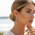 Portrait de profil d'une personne portant une boucle d'oreille pendante dorée ornée de perles de lapis-lazuli bleu intense. Le bijou, composé d'une alternance de pierres bleues et de perles d'or géométriques, ressort avec éclat sur le cou. La personne a les cheveux coiffés en un chignon bas, mettant en lumière le contraste entre le bleu profond des pierres et le teint lumineux.