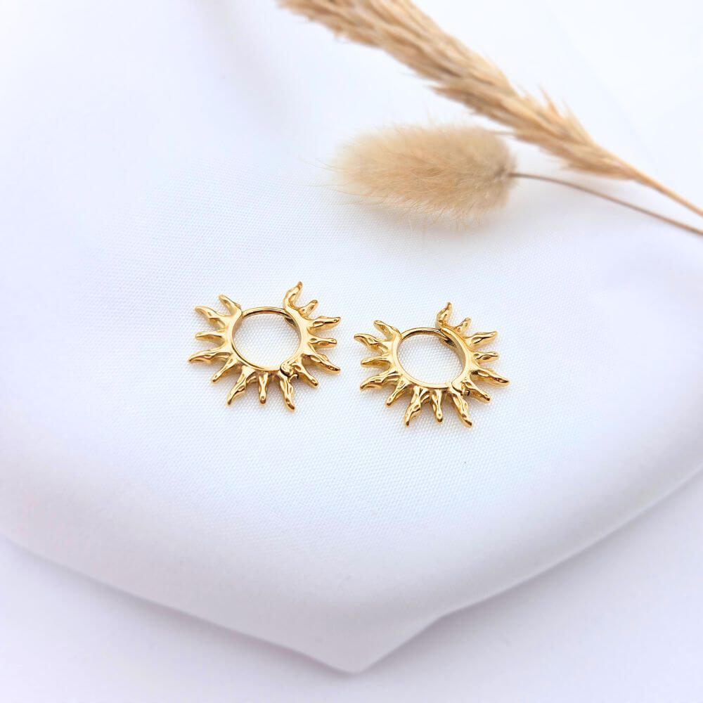 Boucles d'oreilles HELIOS en acier doré avec pendentif médaillon ciselé aux motifs solaires, posées sur un tissu blanc avec un épi de blé.