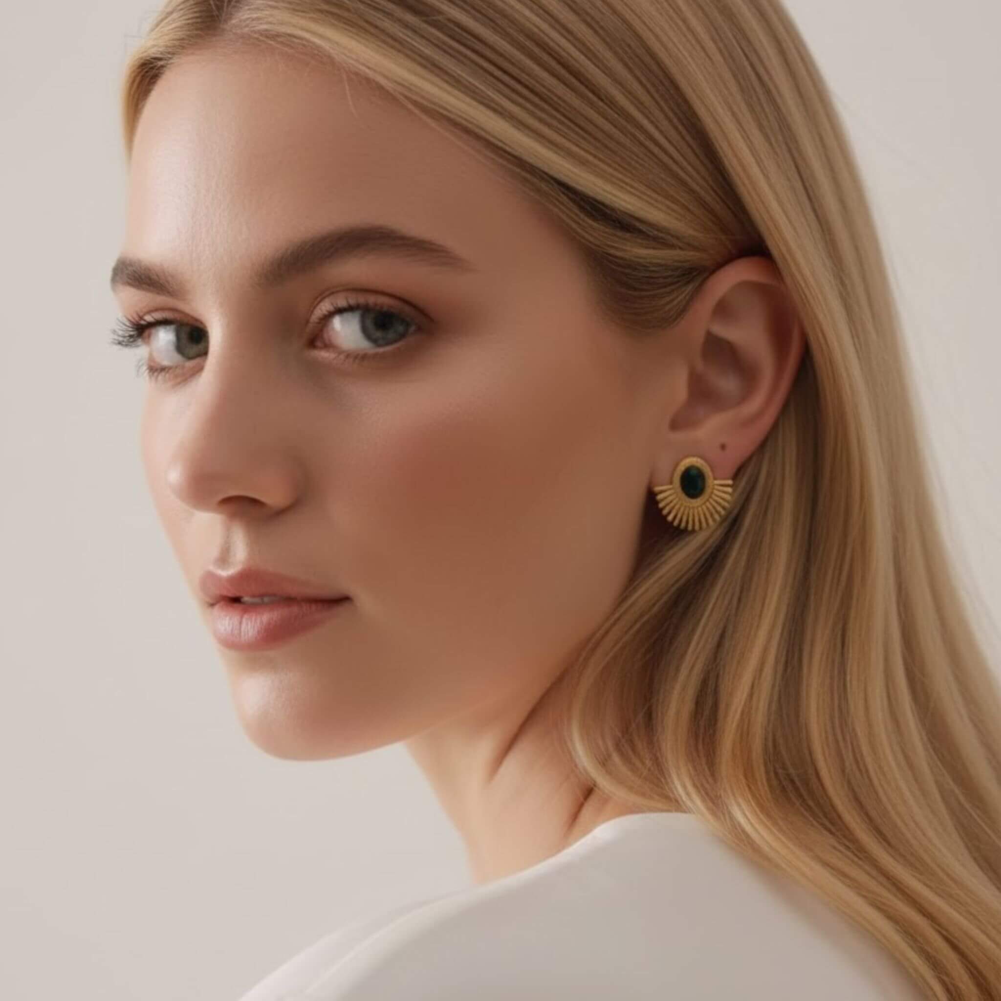Portrait de profil d'une jeune femme blonde portant une boucle d'oreille "soleil" en métal doré. Le bijou présente un design en éventail avec des rayons ciselés entourant une petite pierre ronde d'un vert très foncé. Elle porte un vêtement blanc, sur un fond neutre clair et lumineux.