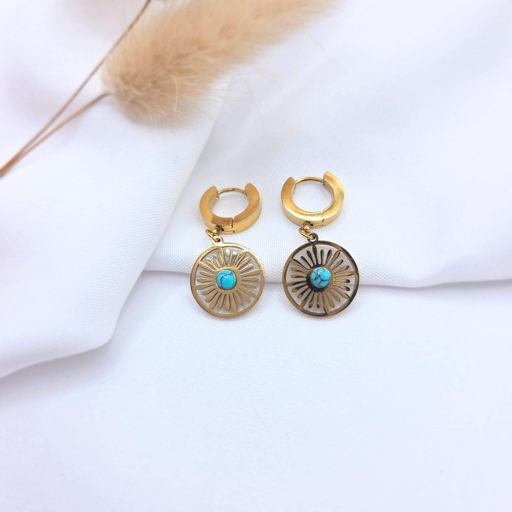 Paire de boucles d'oreilles dormeuses dorées avec pendentifs circulaires ajourés en forme de soleil. Chaque disque présente un centre serti d'une pierre turquoise marbrée. Les bijoux sont posés sur un tissu blanc avec un brin de pampa séchée en arrière-plan.