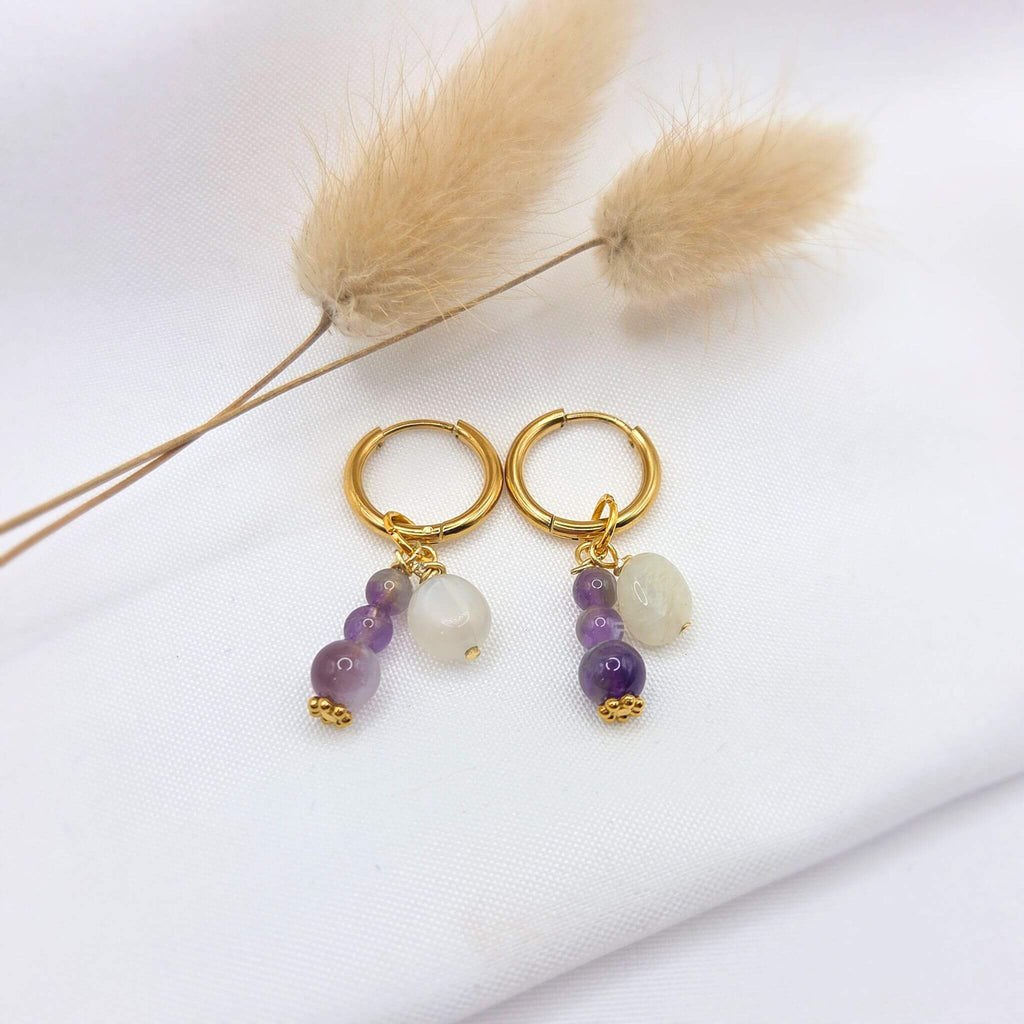Paire de boucles d'oreilles créoles dorées avec pampilles, présentées sur un tissu blanc avec des fleurs séchées. Chaque créole suspend une grappe composée d'une pierre de lune blanche et de trois perles d'améthyste violettes de tailles dégressives, terminées par une petite fleur dorée ciselée.