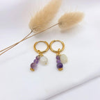 Paire de boucles d'oreilles créoles dorées avec pampilles, présentées sur un tissu blanc avec des fleurs séchées. Chaque créole suspend une grappe composée d'une pierre de lune blanche et de trois perles d'améthyste violettes de tailles dégressives, terminées par une petite fleur dorée ciselée.