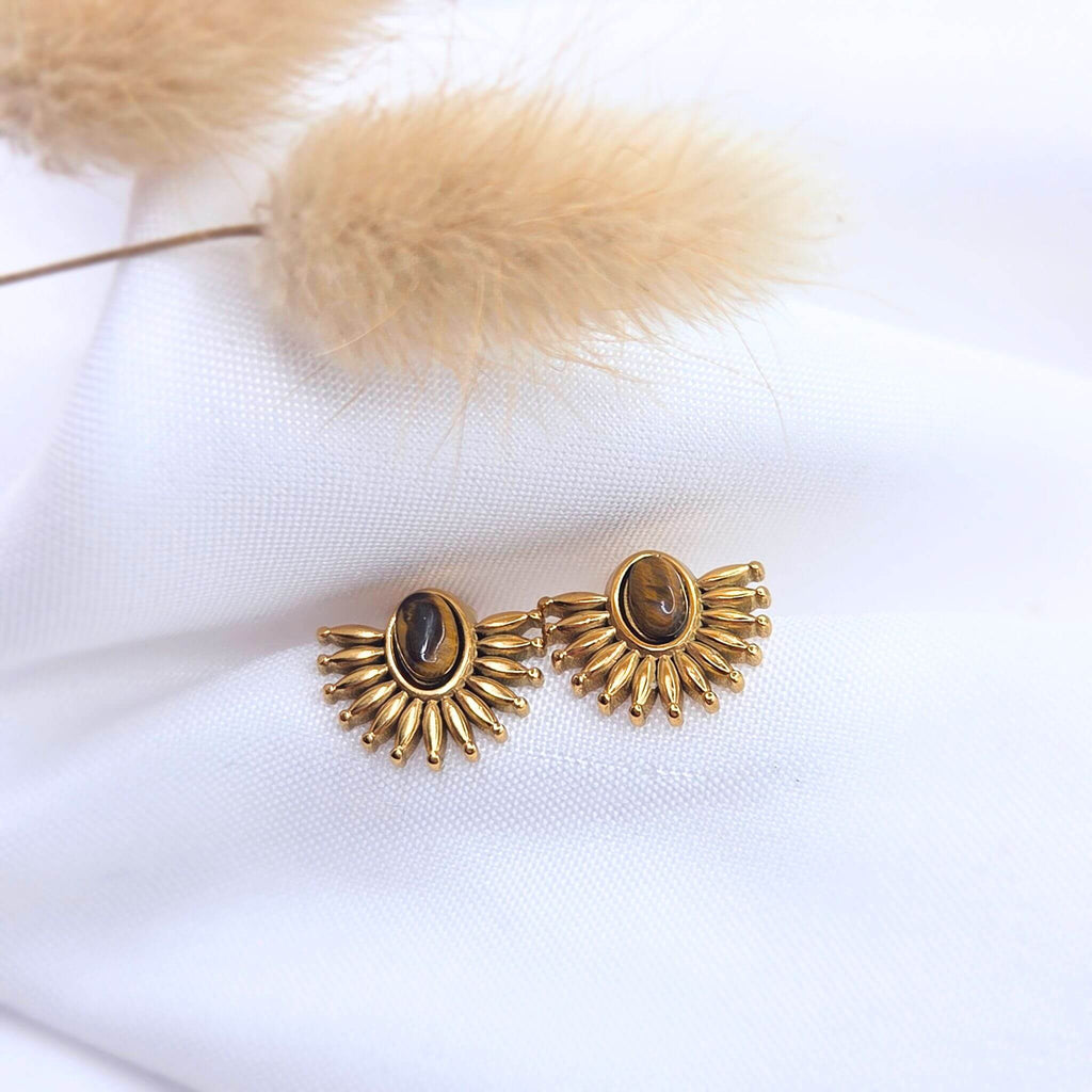 Paire de boucles d'oreilles puces de style éventail ou soleil en métal doré, ornées chacune d'une pierre naturelle œil-de-tigre ovale au centre. Les boucles sont posées sur un tissu blanc texturé à côté d'une fleur de pampa séchée.