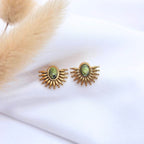 Paire de boucles d'oreilles puces dorées en forme d'éventail ou de soleil levant. Chaque boucle présente une pierre de turquoise africaine ovale d'un vert marbré au centre, entourée de rayons métalliques arrondis. Les bijoux sont posés sur un tissu blanc texturé, à côté d'un plumeau de pampa flou en arrière-plan.