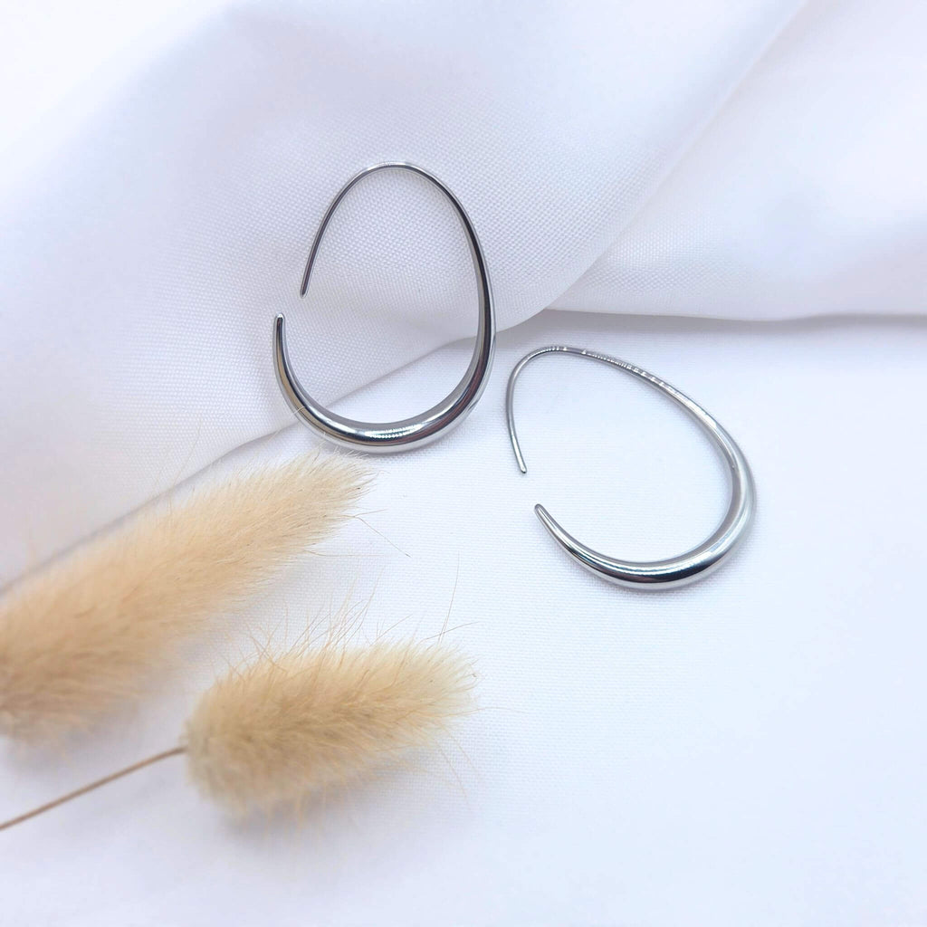 Paire de boucles d'oreilles créoles ovales en argent poli au design minimaliste. Les anneaux présentent une forme de goutte étirée avec une épaisseur progressive sur le bas. Elles sont disposées sur un tissu blanc texturé à côté de fleurs de pampa séchées.