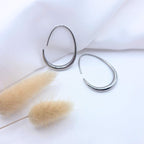 Paire de boucles d'oreilles créoles ovales en argent poli au design minimaliste. Les anneaux présentent une forme de goutte étirée avec une épaisseur progressive sur le bas. Elles sont disposées sur un tissu blanc texturé à côté de fleurs de pampa séchées.