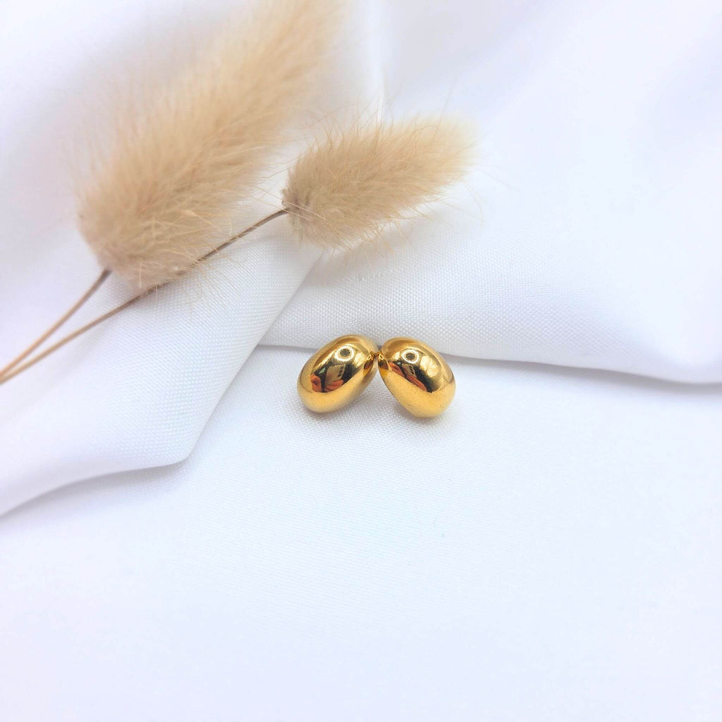 Paire de boucles d'oreilles clous de forme ovale et bombée en métal doré brillant, posée sur un tissu blanc drapé. En arrière-plan, deux tiges d'herbe de la pampa séchée complètent cette composition élégante et lumineuse.