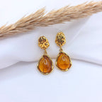 Paire de boucles d'oreilles pendantes dorées composées d'un clou texturé de forme organique et d'une pierre ambrée en forme de goutte. Les pierres translucides aux reflets miel sont serties de métal doré brillant, posées sur un tissu blanc avec un épi de blé séché en arrière-plan.