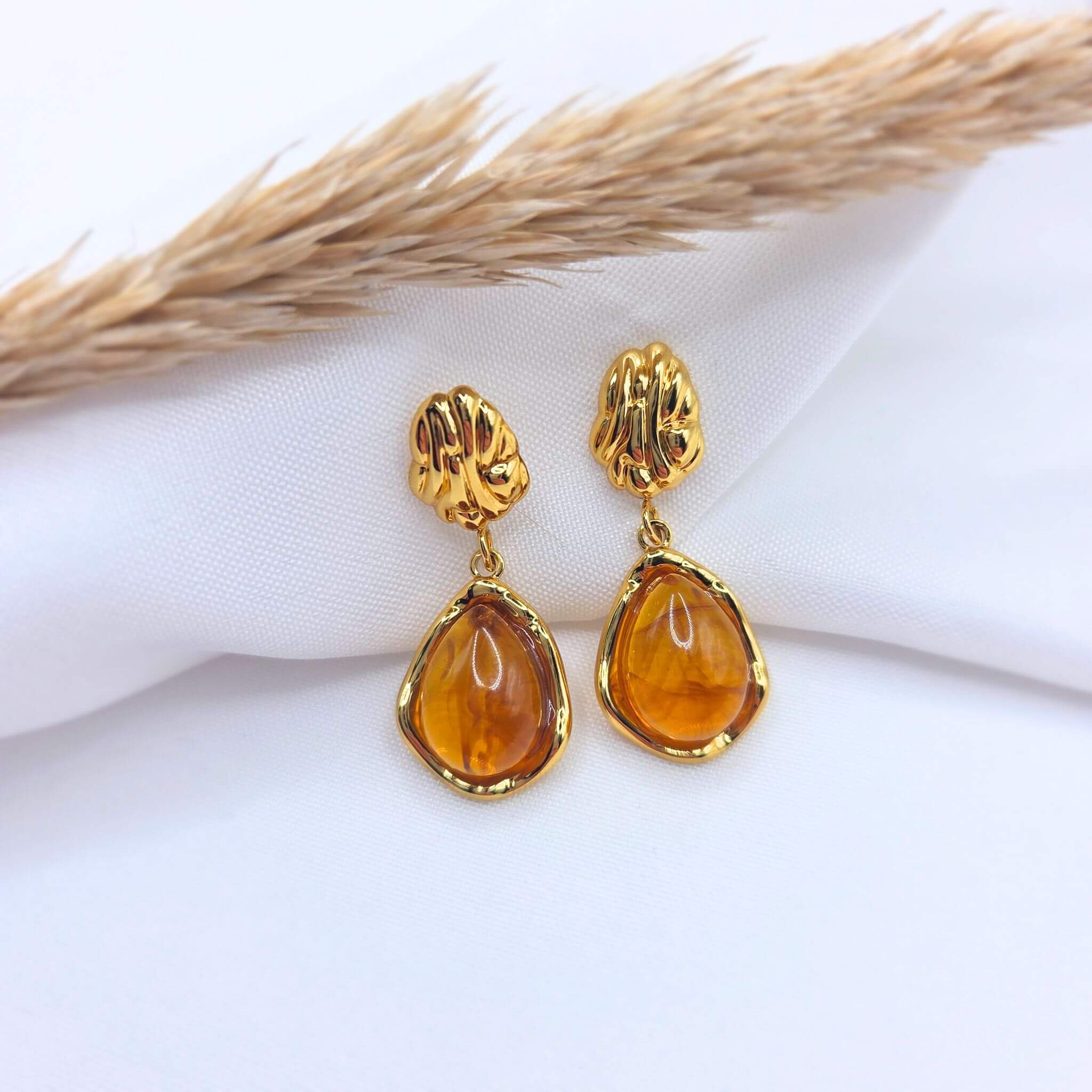 Paire de boucles d'oreilles pendantes dorées composées d'un clou texturé de forme organique et d'une pierre ambrée en forme de goutte. Les pierres translucides aux reflets miel sont serties de métal doré brillant, posées sur un tissu blanc avec un épi de blé séché en arrière-plan.