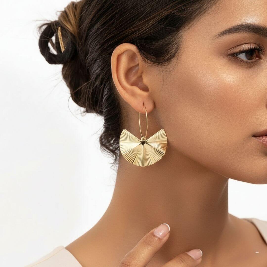 Portrait de profil d'une femme portant une boucle d'oreille dorée en forme d'éventail plissé. La créole est fine et le pendentif texturé capte la lumière. Ses cheveux sont attachés en un chignon flou, mettant en valeur le bijou.