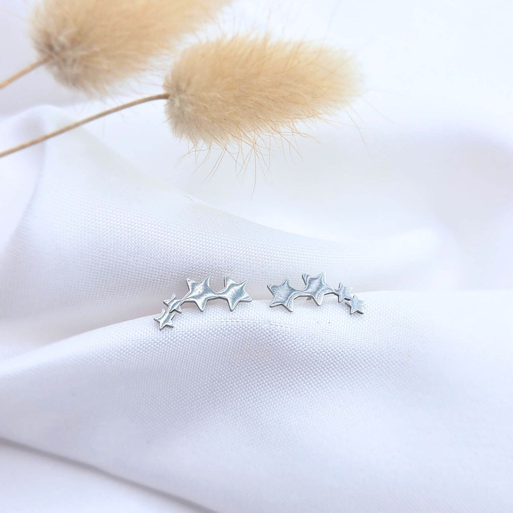 Boucles d'oreilles 'Ear Crawler' étoiles en acier inoxydable argenté. Design céleste et minimaliste présenté dans un décor épuré avec drapé blanc et pampa.