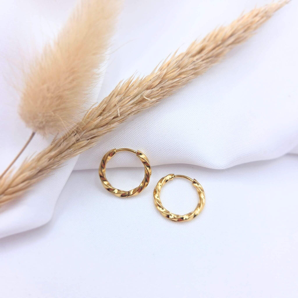 Paire de petites boucles d'oreilles créoles dorées au design torsadé. Les anneaux présentent une finition brillante et texturée en spirale, posés sur un tissu blanc drapé à côté d'un épi de blé et de fleurs de pampa séchées.