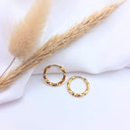 Paire de petites boucles d'oreilles créoles dorées au design torsadé. Les anneaux présentent une finition brillante et texturée en spirale, posés sur un tissu blanc drapé à côté d'un épi de blé et de fleurs de pampa séchées.
