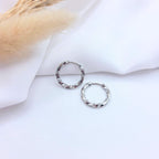 Paire de petites boucles d'oreilles créoles en argent au design torsadé. Les anneaux présentent un effet de spirale poli qui accroche la lumière, posés sur un tissu blanc drapé à côté de fleurs de pampa séchées.