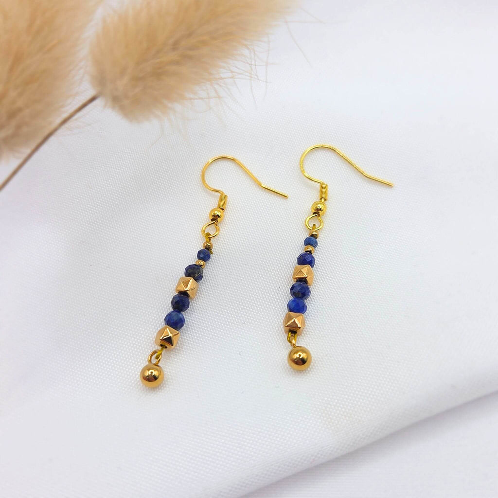 Paire de boucles d'oreilles pendantes dorées ornées de perles de lapis-lazuli bleu profond facettées. Les pierres sont alternées avec des perles d'or aux formes géométriques (cubes et sphères), créant une ligne verticale élégante. Les bijoux sont disposés sur un tissu blanc avec un détail de fleur séchée en arrière-plan.