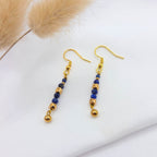 Paire de boucles d'oreilles pendantes dorées ornées de perles de lapis-lazuli bleu profond facettées. Les pierres sont alternées avec des perles d'or aux formes géométriques (cubes et sphères), créant une ligne verticale élégante. Les bijoux sont disposés sur un tissu blanc avec un détail de fleur séchée en arrière-plan.