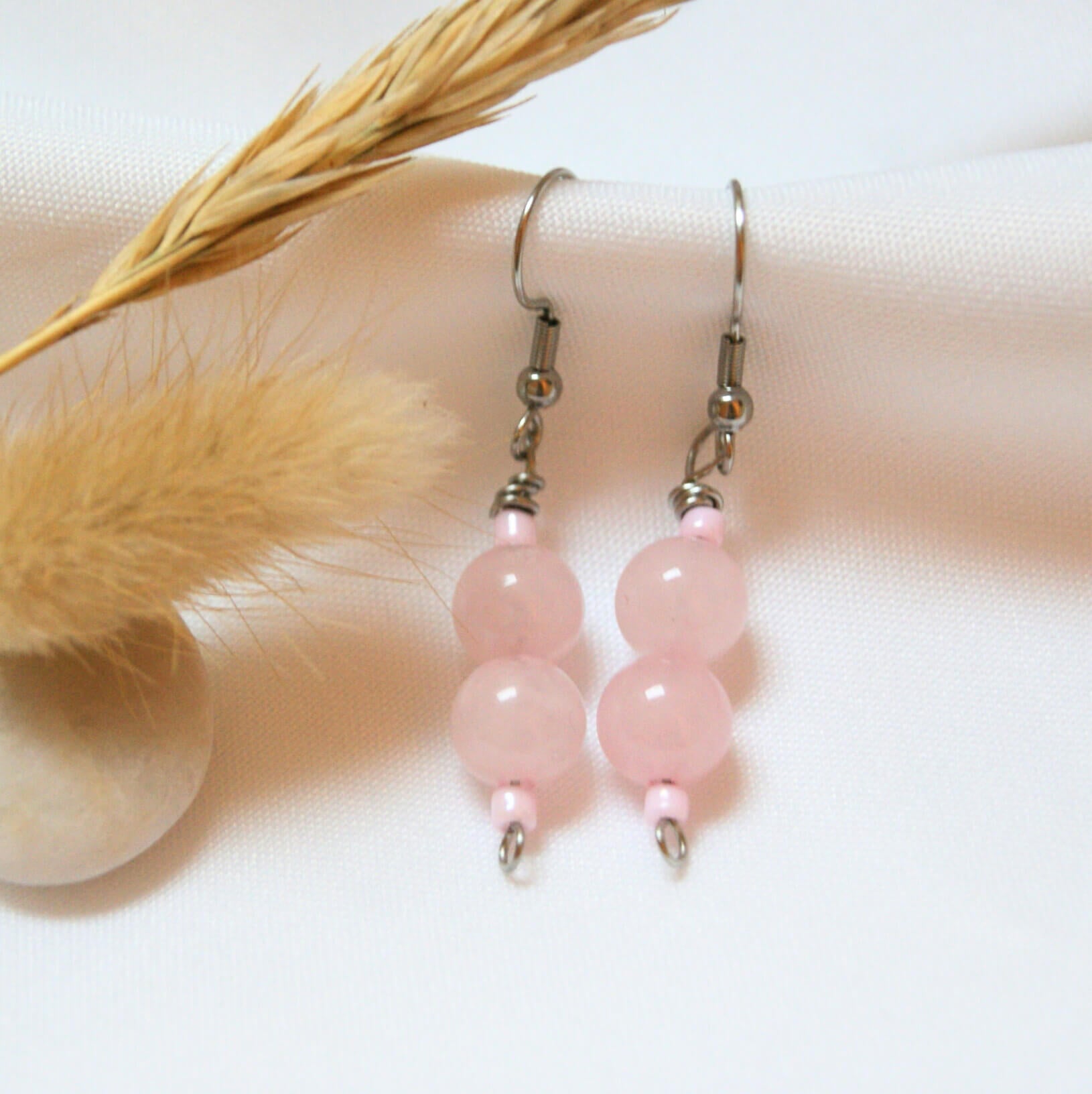 Une paire de boucles d'oreilles pendantes délicates, chacune ornée de deux perles rondes en quartz rose translucide. Les perles sont suspendues à des crochets argentés et séparées par de petites perles de rocaille rose pâle. Le bijou est présenté sur un fond de tissu blanc texturé, accompagné d'un épi de blé séché et d'un plumeau végétal beige, créant une atmosphère douce et bohème.