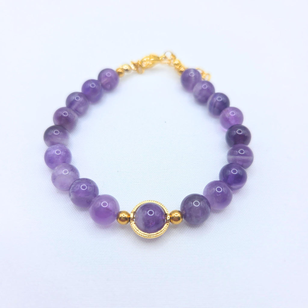 Bracelet en perles d'améthyste naturelle violette avec un médaillon central doré et un fermoir mousqueton en or, présenté sur un fond blanc.
