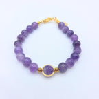Bracelet en perles d'améthyste naturelle violette avec un médaillon central doré et un fermoir mousqueton en or, présenté sur un fond blanc.