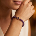Un gros plan de face montre une femme, du bas du visage jusqu'au coude, posant son menton sur le dos de sa main droite pliée. Sur son poignet se trouve un bracelet "Nora" en perles d'améthyste violettes et rondes. Au centre du bracelet se trouve une seule perle d'améthyste violette sertie d'un anneau en acier inoxydable doré, encadré de chaque côté par une petite perle dorée. La femme porte une robe de couleur crème, et l'arrière-plan est flou, dans des tons beiges.