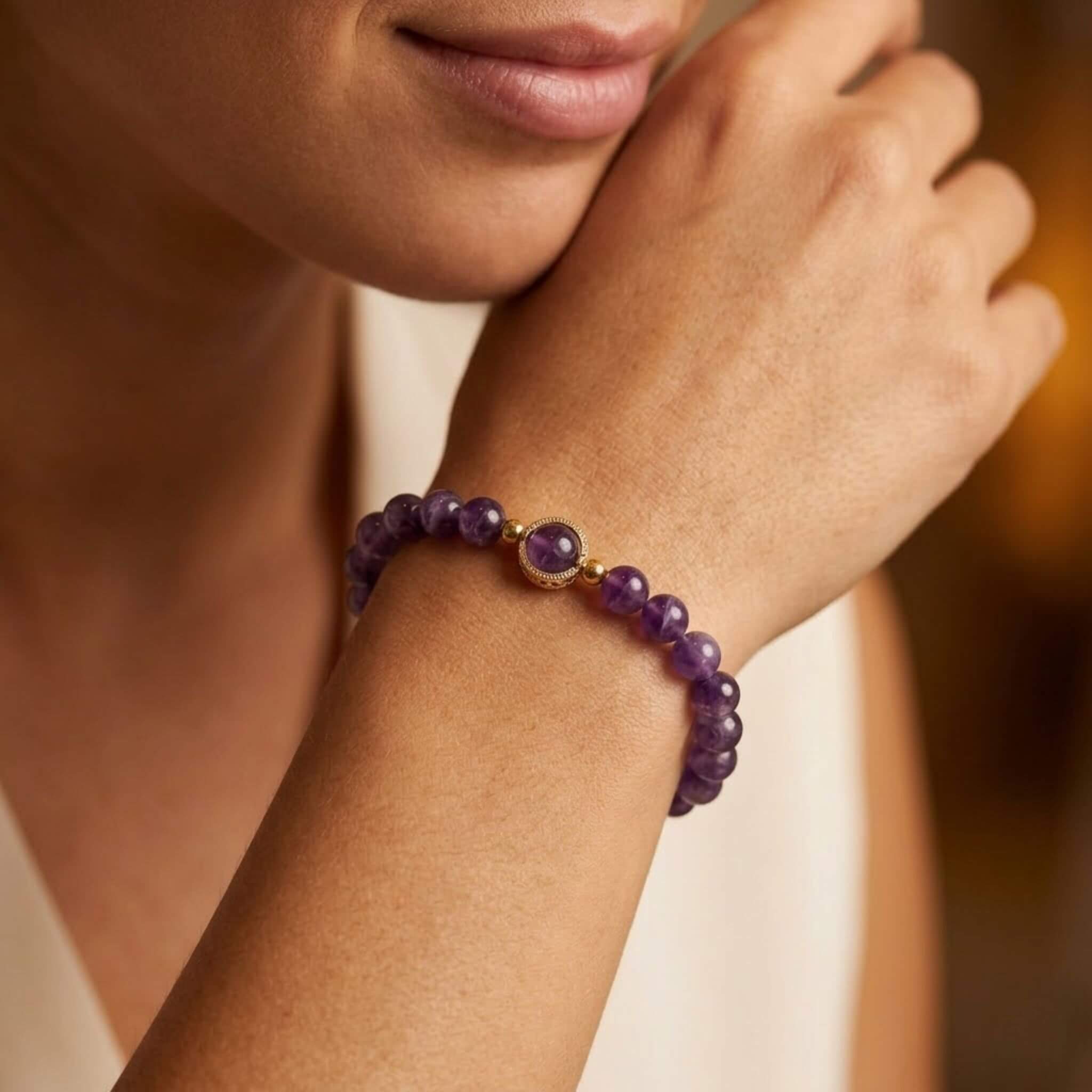 Un gros plan de face montre une femme, du bas du visage jusqu'au coude, posant son menton sur le dos de sa main droite pliée. Sur son poignet se trouve un bracelet "Nora" en perles d'améthyste violettes et rondes. Au centre du bracelet se trouve une seule perle d'améthyste violette sertie d'un anneau en acier inoxydable doré, encadré de chaque côté par une petite perle dorée. La femme porte une robe de couleur crème, et l'arrière-plan est flou, dans des tons beiges.