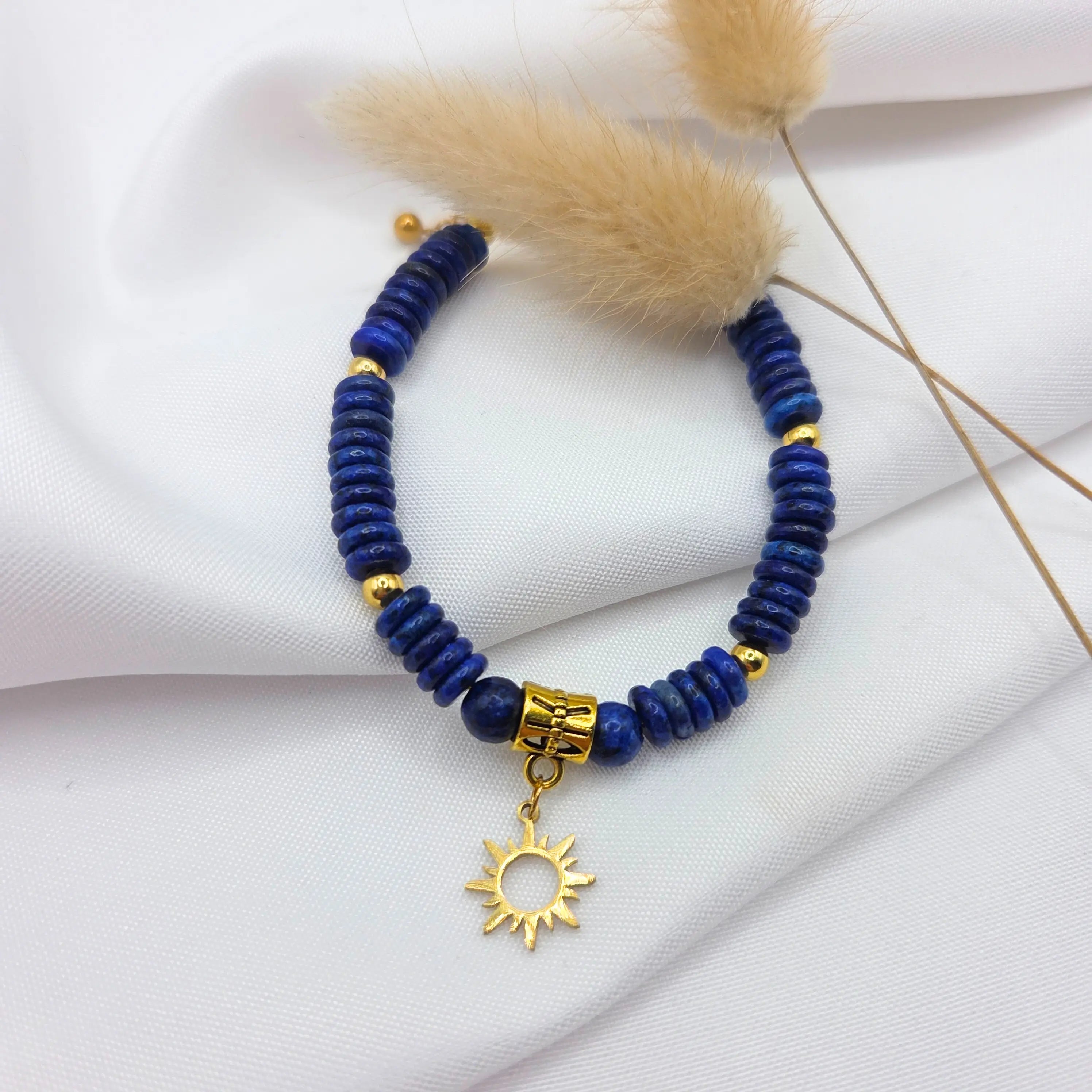 Vue de dessus d'un bracelet composé de perles rondelles en pierre bleu foncé intense de Lapis Lazuli sur un tissu blanc drapé. Le bijou est ponctué de quelques billes dorées et présente en son centre un pendentif en forme de soleil doré aux rayons ciselés. Deux plumeaux de graminées séchées sont disposés en arrière-plan pour la décoration.